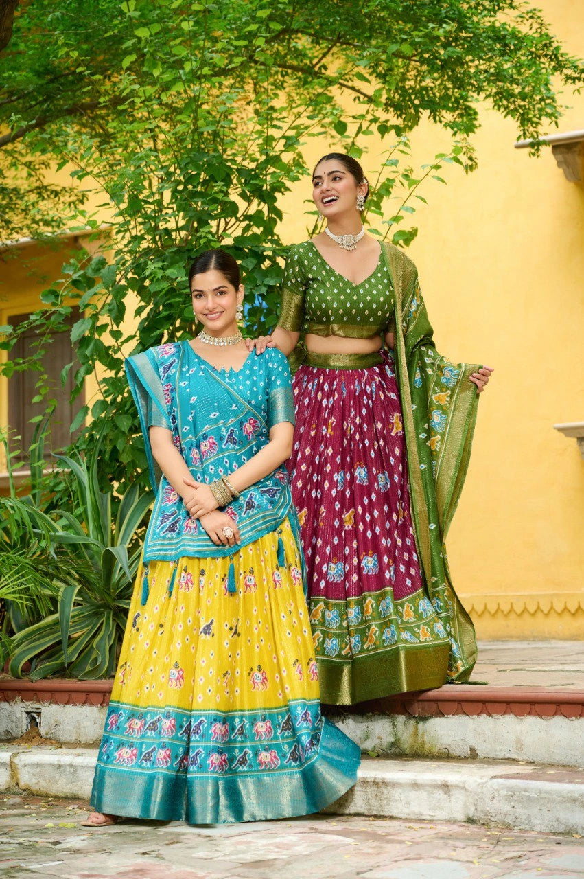 Two women in colorful traditional outfits standing outdoors with a yellow wall and greenery in the background.